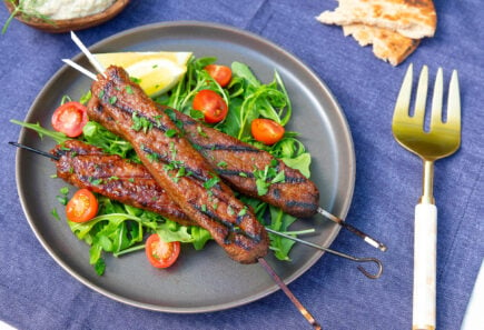 A plate piled with grilled beyond meat plant-based mediterranean skewers on a bed of greens and grape tomatoes