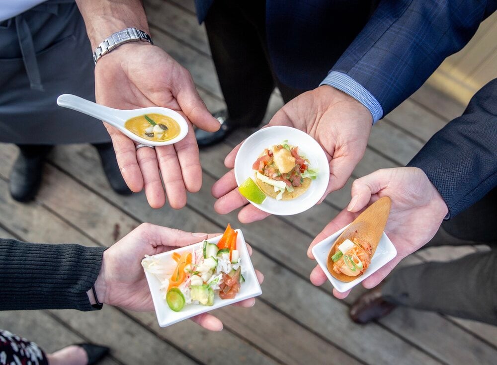 A close up on the hands of four people in business attire holding bluenalu cultivated seafood-based appetizers on small plates