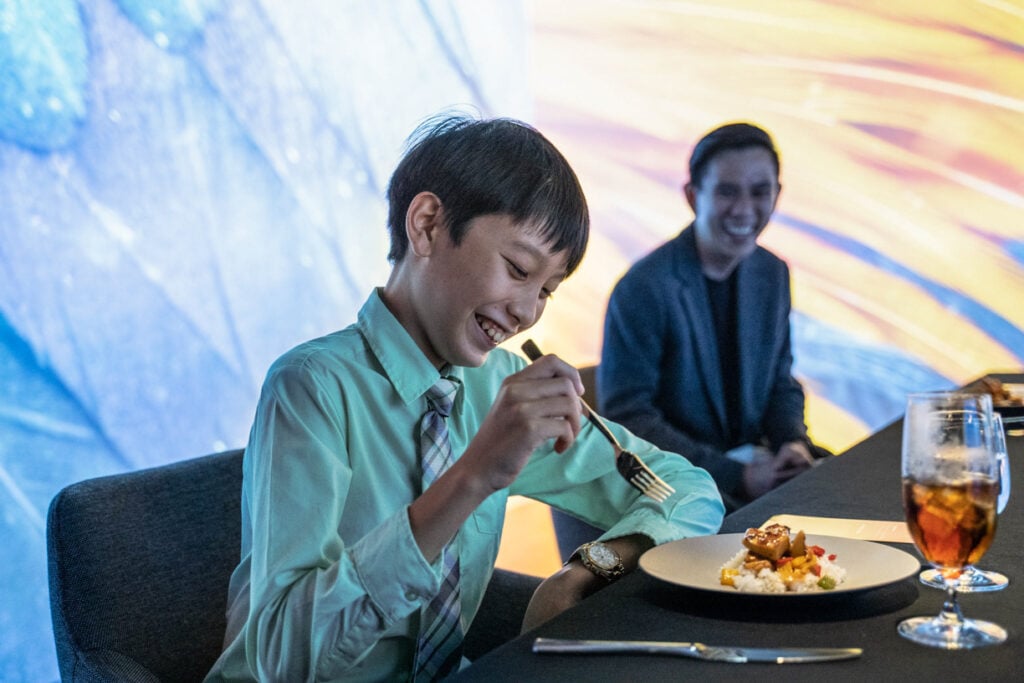Boy eating food with person laughing behind