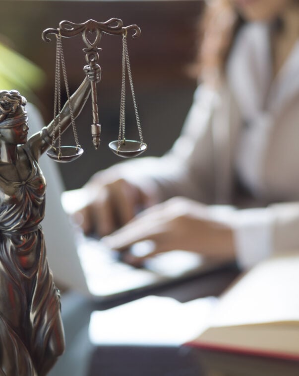 A small figurine of the scales of justice in front of a woman working on her laptop