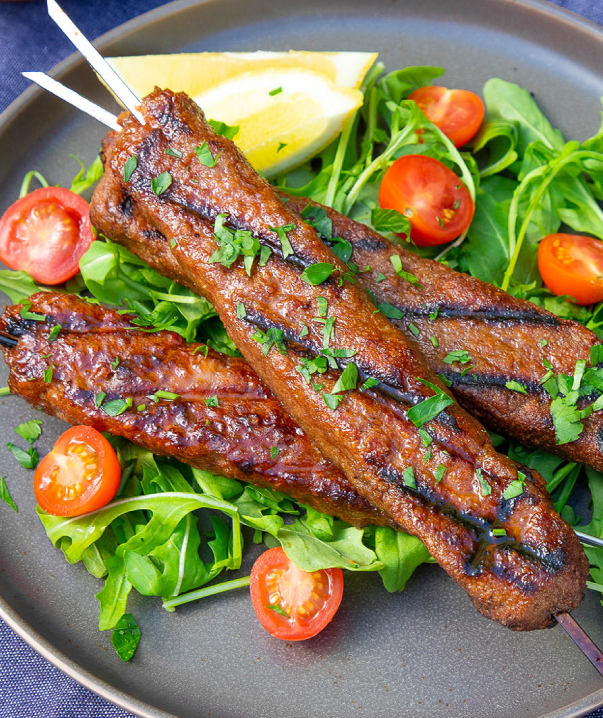 A plate piled with grilled beyond meat plant-based meditteranean skewers on a bed of greens and grape tomatoes